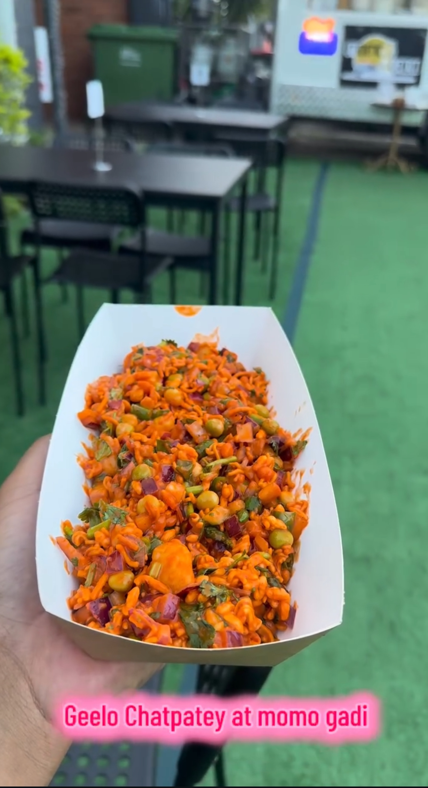 A takeaway tray of Geelo Chatpatey held up beside the outdoor tables
